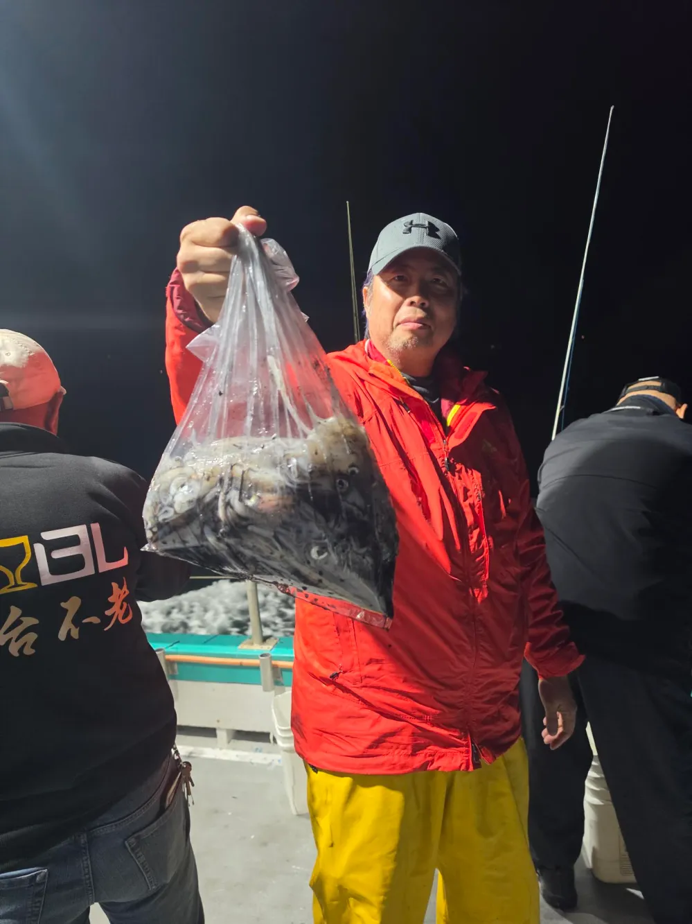 Man in red jacket holding a bag of fish on a boat at night.