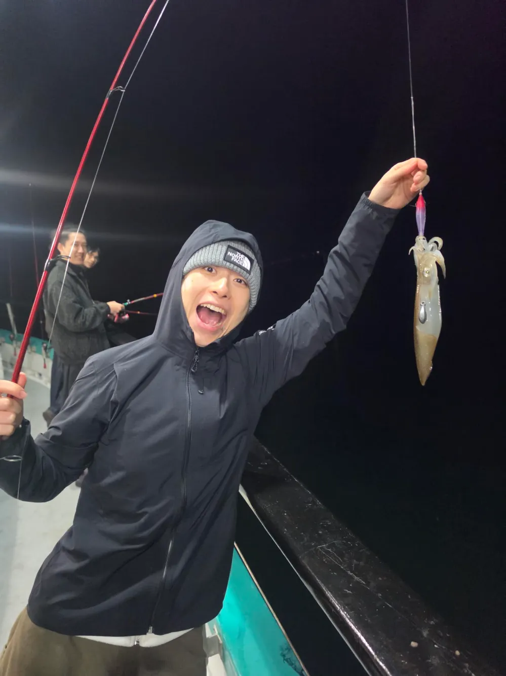 Person excitedly holding a fishing rod with a squid, wearing a beanie and jacket at night on a boat.