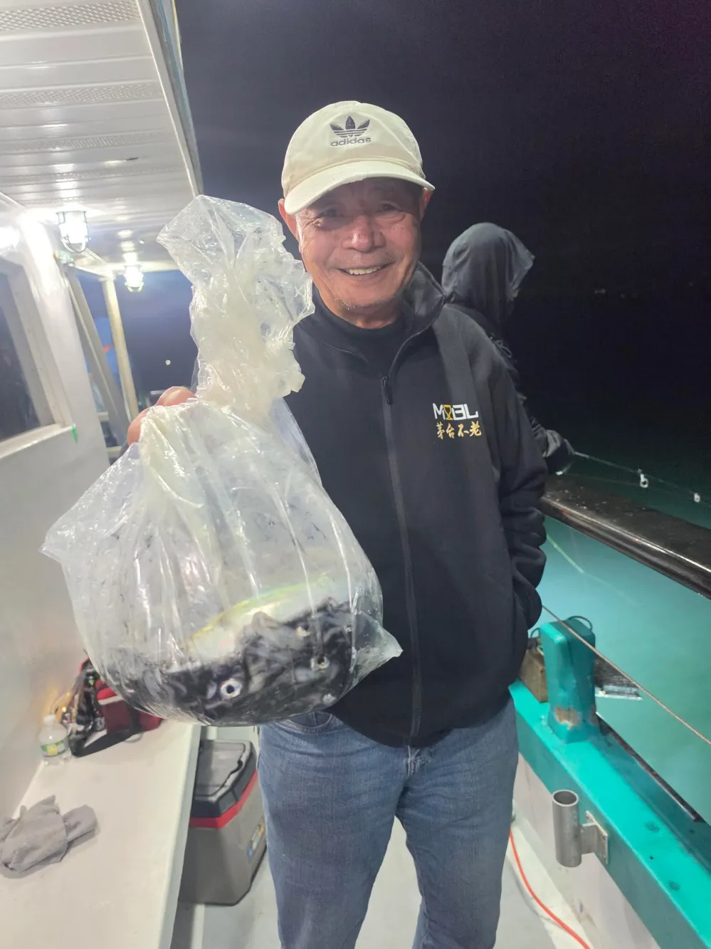 Man holding a bag of fish on a boat at night, smiling.
