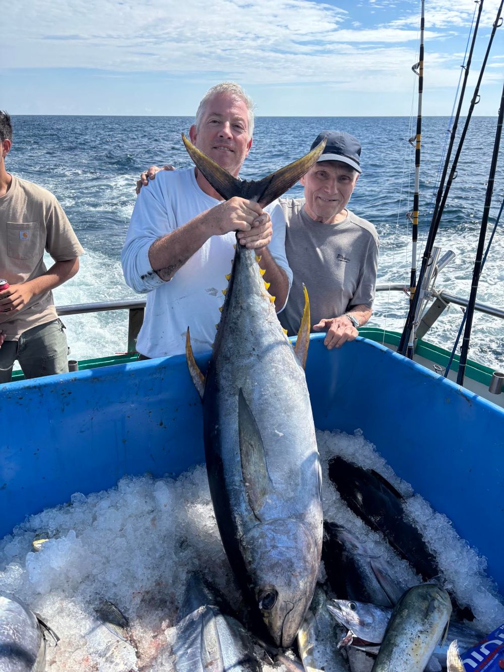 Two people on a boat holding a large tuna, surrounded by a bin of fish on ice.
