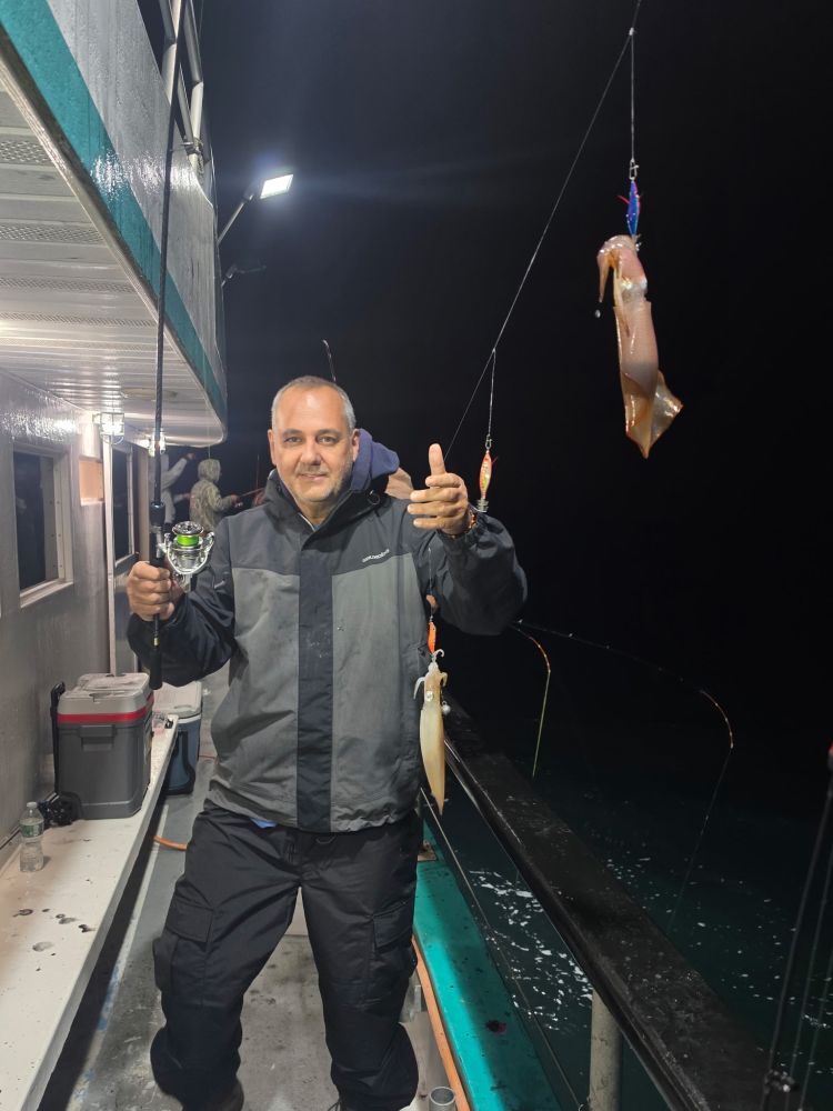 Man on a boat holding a fishing rod with two squids at night.