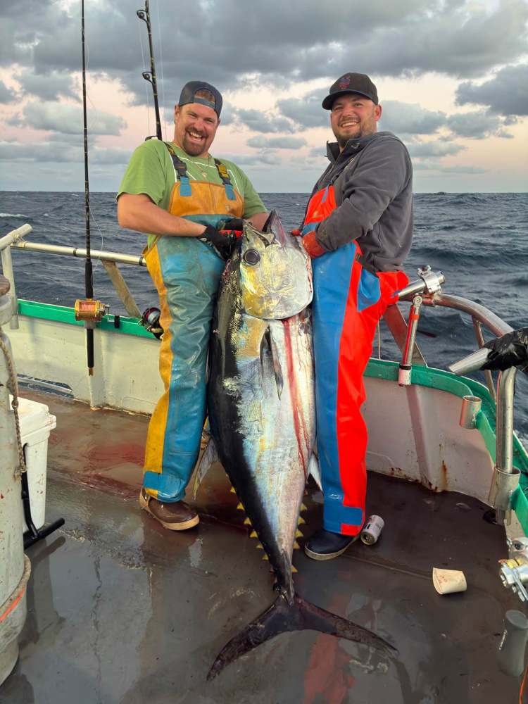 Two smiling fishermen on a boat hold a large tuna.