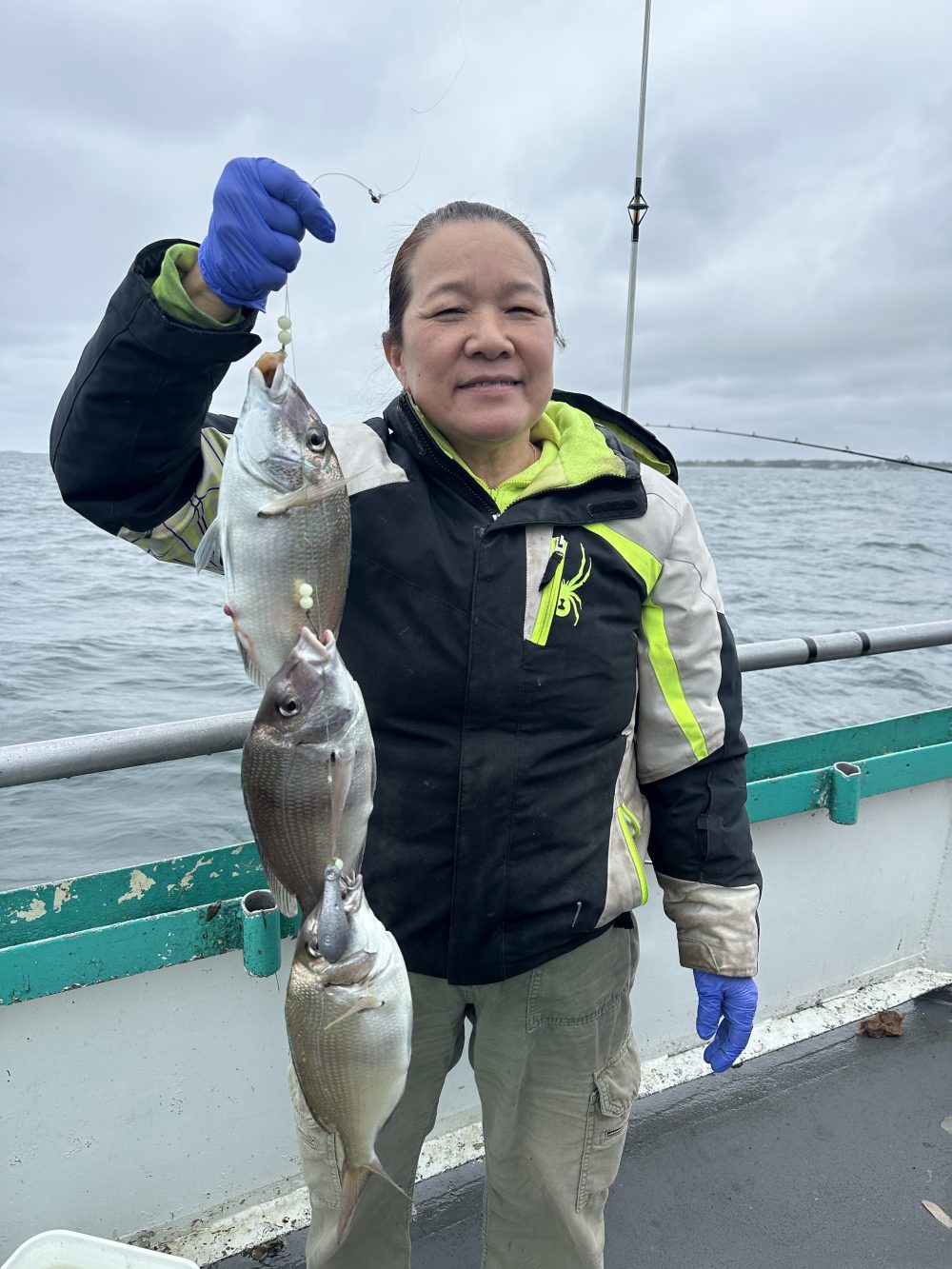 Person on boat holding two fish, wearing gloves and a jacket, with ocean in the background.