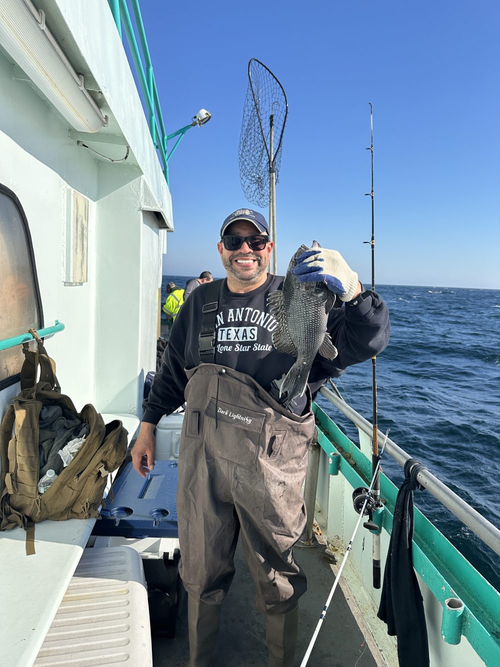 Person holding a fish on a boat, wearing waders and a hat, with fishing gear visible.