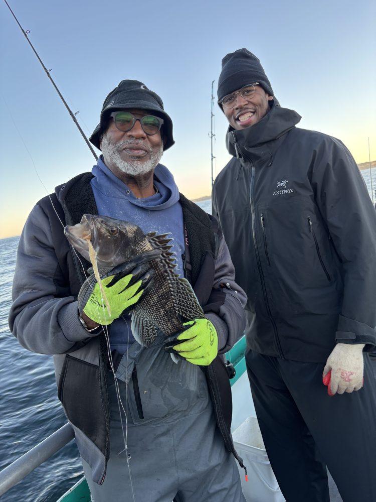 Two people on a boat, one holding a fish, both wearing jackets and hats.