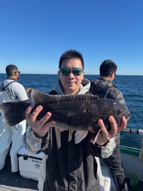 Person holding a large fish on a boat with ocean and people in background.