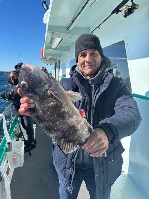Person holding a large fish on a boat, wearing a dark jacket and beanie.